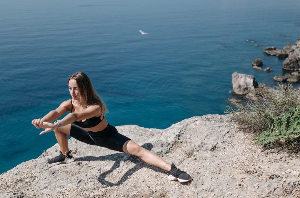 A woman in activewear performs stretches on a cliffside overlooking the ocean on a sunny day.