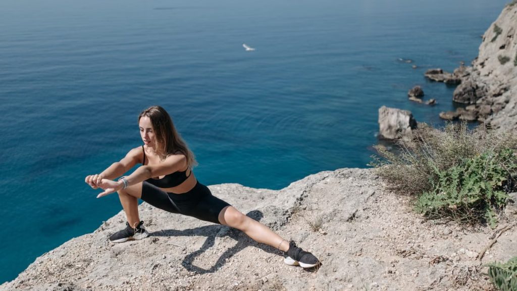 A woman in activewear performs stretches on a cliffside overlooking the ocean on a sunny day.