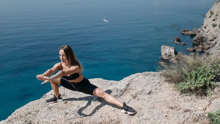 A woman in activewear performs stretches on a cliffside overlooking the ocean on a sunny day.