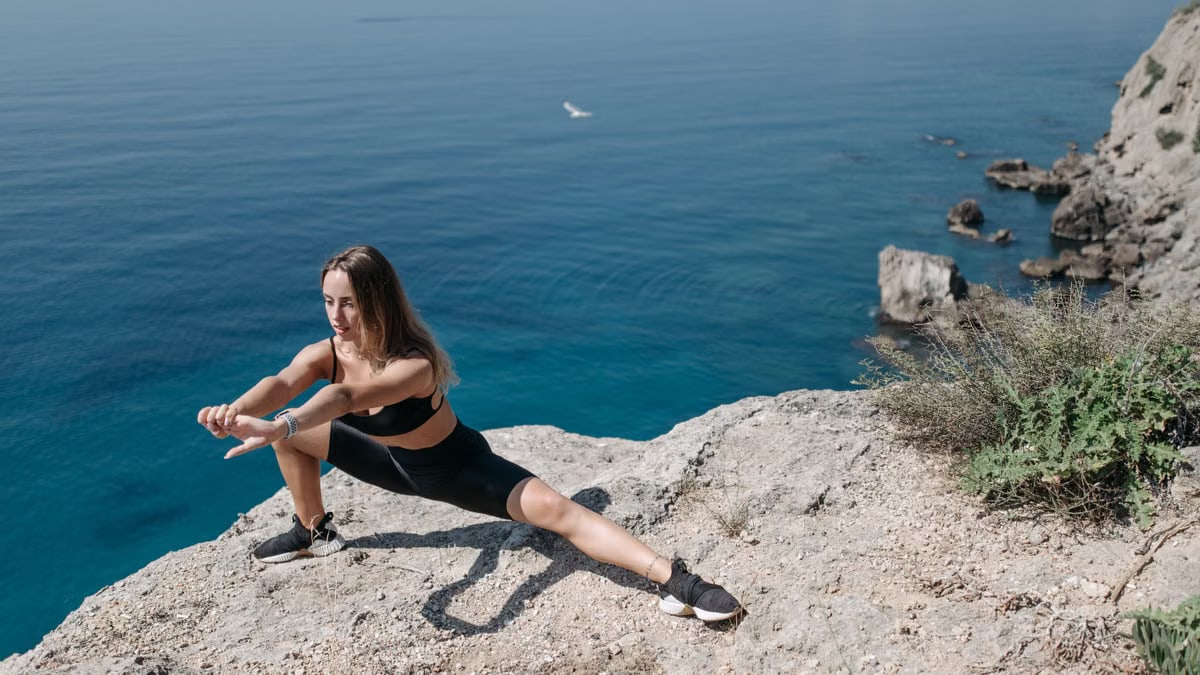 A woman in activewear performs stretches on a cliffside overlooking the ocean on a sunny day.